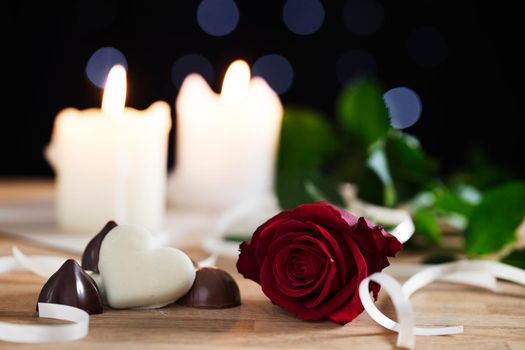 Red rose on table with chocolates and candles in background, shallow depth of field.