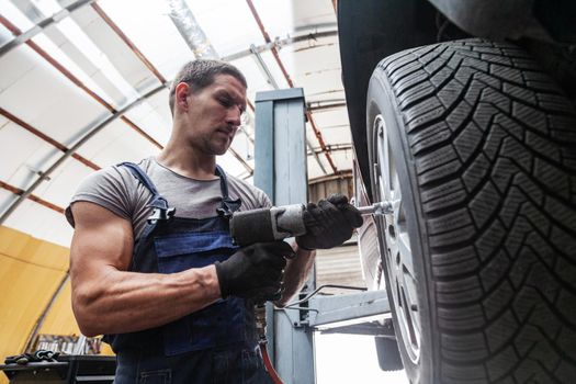 A muscular mechanic is maintaining wheels on a vehicle in auto garage with pneumatic tool.
