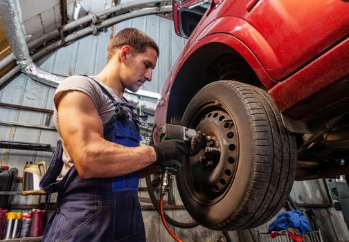 A muscular mechanic is maintaining wheels on a vehicle in auto garage with pneumatic tool.