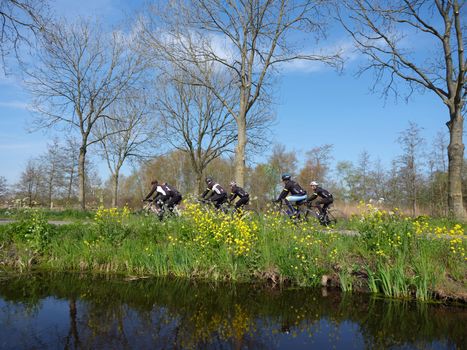 group of men on racing bikes on country road in holland near woerden under blue sky in spring with yellow flowers and canal