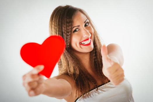 Close-up of a beautiful smiling girl holding a red heart with tumb up. Selective focus. Focus on the girl.