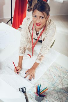 Young smiling female fashion designer with tape measure draped over the neck drawing on sewing pattern with a ruler. Looking at camera. Top view.