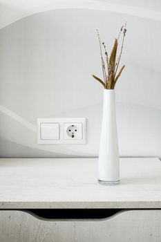 Stylish wooden bedside table and white glass vase with dried branch and plants on it. Interior details. Vertical image.