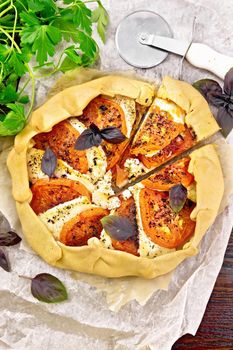 Pie with tomatoes, curd cheese and purple basil on parchment, parsley on a wooden board background from above