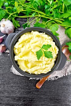 Mashed potatoes in a black saucepan and a spoon on burlap, garlic, parsley on dark wooden board background from above