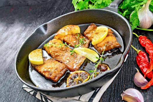 Pink salmon with a sauce of honey, lemon juice, garlic, hot pepper and soy sauce, lemon slices and sprig of thyme in a frying pan on a towel, parsley on black wooden background
