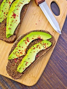 Two slices of rye bread with slices of avocado and pepper, knife against the dark wooden board