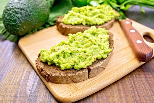 Two slices of rye bread with guakomole avocado on a plate, napkin, parsley on a wooden boards background