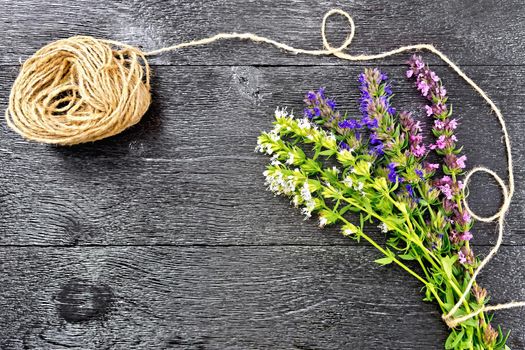 Bunch of fresh savory with green leaves and flowers, a coil of twine against a black wooden board