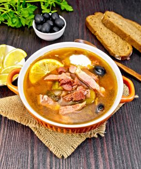 Soup saltwort with lemon, meat, pickles, tomato sauce and olives in a sackcloth, bread and parsley on a dark wooden board