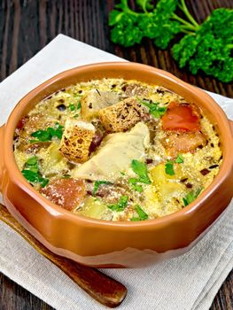 Fish soup with tomatoes, potatoes, peppers and cream in a clay bowl on a napkin, parsley, spoon on a wooden board background