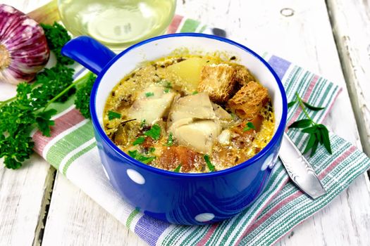 Fish soup with tomatoes, potatoes, peppers, croutons and cream in a blue bowl on a napkin, parsley and garlic on a light wooden board background