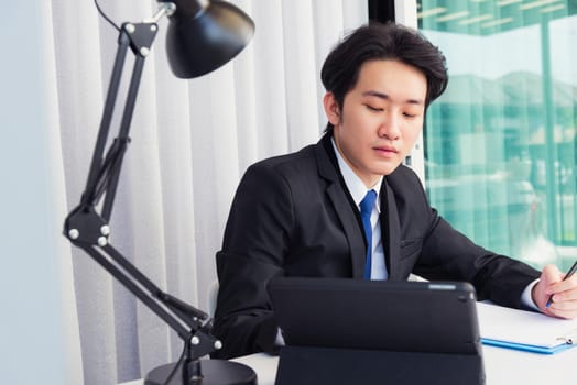 Work from home, Asian young businessman video conference call or facetime he smiling looking to tablet sitting on desk using smart digital tablet computer and write notebook information at home office