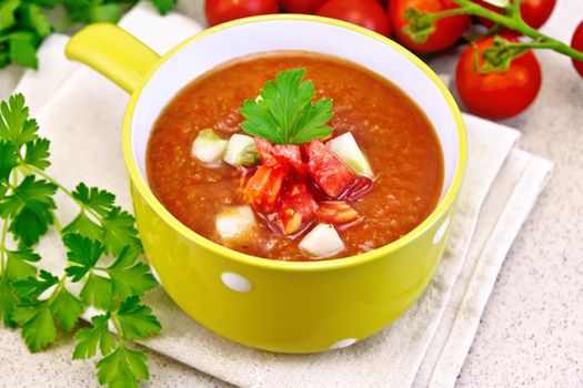 Gazpacho tomato soup in yellow bowl with parsley and vegetables on a towel on the background of a granite table