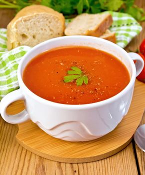 Tomato soup in a white bowl, napkin, spoon, tomato, parsley, bread on a wooden boards background