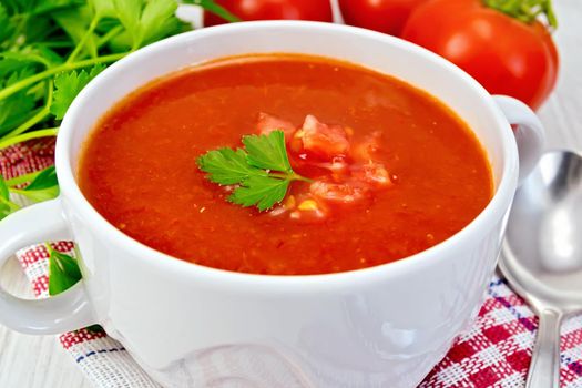 Tomato soup with chunks of vegetable in a white bowl on a napkin with a spoon, tomatoes, parsley on a lighter background board