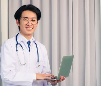 Portrait of Asian young handsome doctor man wearing a doctor's dress and stethoscope smiling sitting at hospital office, Health medical care concept