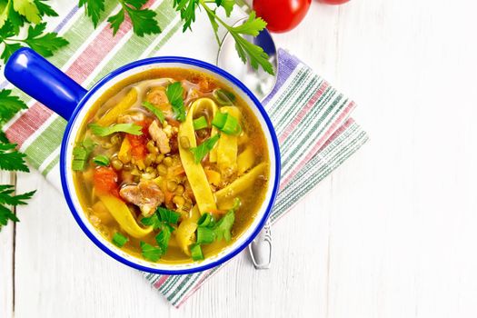 Soup with meat, tomatoes, vegetables, mung bean lentils and noodles in a blue bowl on a towel, parsley and spoon on wooden board background from above