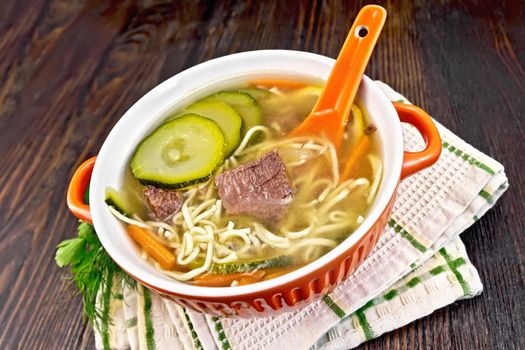 Soup with zucchini, beef, ham, lemon and noodles in a red bowl, parsley and dill on a kitchen towel against a dark wooden board