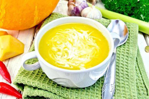 Soup-puree pumpkin with cream in a white bowl on a kitchen towel with a spoon, pumpkin, hot red pepper pods, leek and garlic, parsley on a wooden boards background