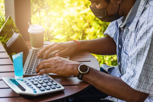 Happy Asian black businessman person or office worker wear face mask protect sitting on desk work from home using laptop computer typing keyboard and connect networking online at the coffee cafe shop