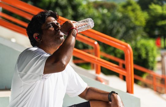 Close up Asian young sport runner black man wear athlete headphones he drinking water from a bottle after running at the outdoor street health park, healthy exercise workout concept