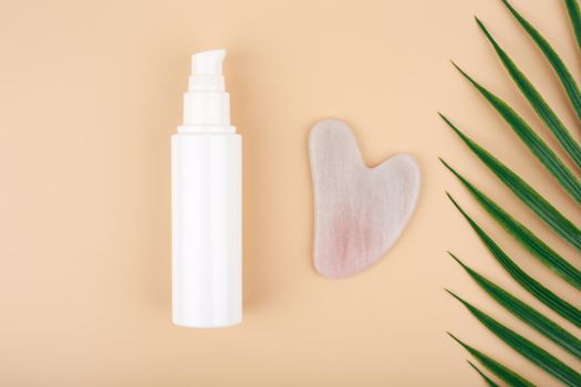 Top view of skin lotion, rose quartz heart shaped guasha massage stone and half of palm leaf on light beige background. Concept of facial massage with guasha scraper