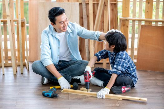 Asian father touch head of his son during drill the timber in home workplace of carpentering with happy emotion. Asian family concept to stay at home and enjoy good relationship hobby together.