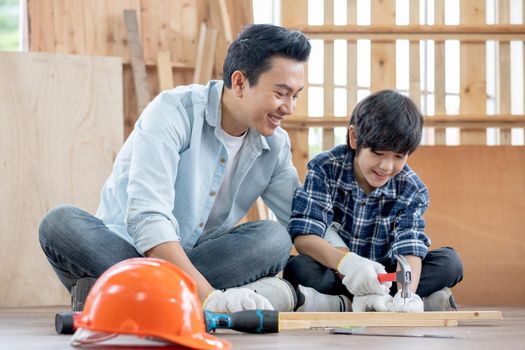 Asian father look carefully son use hammer to hobnailed in their workplace of carpentering with happy emotion. Asian family concept to stay at home and enjoy good relationship hobby together.