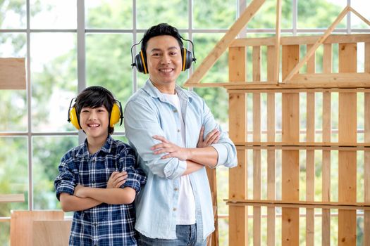 Portrait of Asian father and boy with mechanic headphone stand in front of woodwork and look at camera.