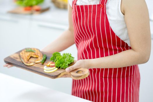 Close up hands of woman hold wooden plate with piece of fish or meat on wood plate stay in kitchen in her house.