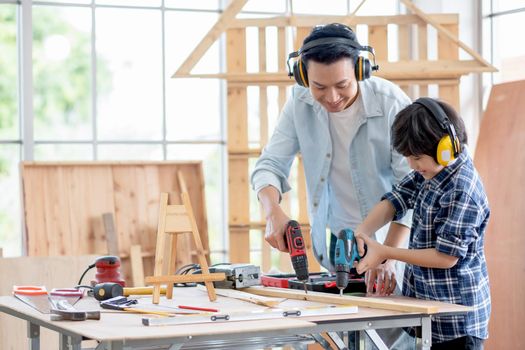 Asian father with noise canceling headphone teach and show to his son to use electric drill to work with woodwork in their house. Concept of good relationship with hobby or activities in happy family.