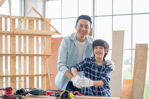 Asian father and boy hold electric drill for woodwork and look to camera with smiling. Concept of good relationship with hobby or activities in happy family.