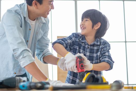 Asian father teach his son to use electric drill to work with woodwork in their house. Concept of good relationship with hobby or activities in happy family.