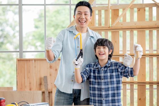 Farther and son express happy and cheerful emotion in room with woodwork and boy hold Hack Saw and piece of wood stand in front of his father. Concept of craftsman family and have good hobby together.