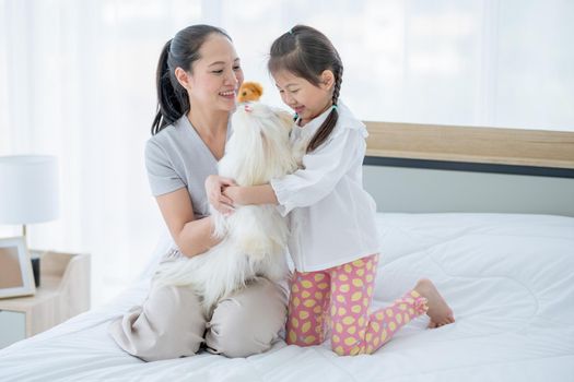 Little Asian girl play with white dog and sit near her mother to take care in bed room. Concept of happy family feel relax to stay home with their own pets.