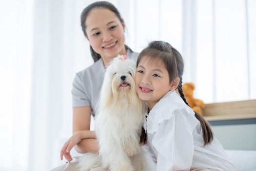 Little Asian girl play with white dog and sit near her mother to take care in bed room. Concept of happy family feel relax to stay home with their own pets.