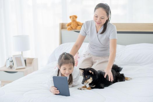 Asian little girl show tablet and make her black Shiba dog interest and stay with mother on white bed. Concept of happy family feel relax to stay home with their own pets.