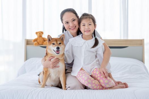 Asian mother and girl sit on white bed with Shiba dog and look to camera with smiling in bedroom. Concept of happy family feel relax to stay home with their own pets.