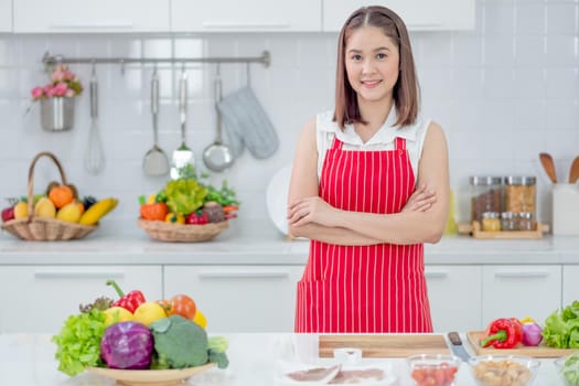 Asian beautiful woman with red apron stand with arm crossed and smiling in kitchen and look to camera with different ingredients and food. Concept of happiness of cooking in their house.