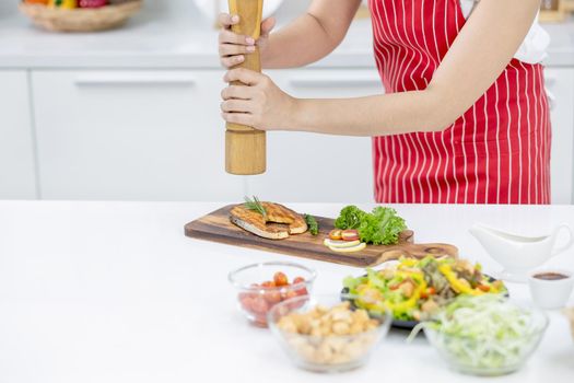 Hands of woman with red apron scatter spices over plate of slicing fish in kitchen with different type of ingredients and garnish on table. Concept of happiness of cooking in their house.