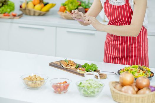 Hands of woman with red apron use mobile phone take photo of plate of slicing fish in kitchen with different type of ingredients and garnish on table. Concept of happiness of cooking in their house.