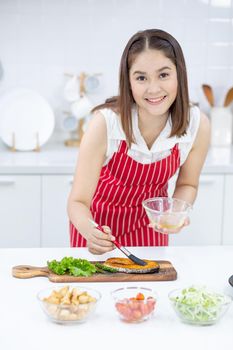 Asian beautiful woman with red apron smear oil and garnish on slicing fish on plate in kitchen with different types of bowl of ingredients on table. Concept of happiness of cooking in their house.