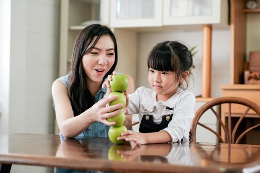 Asian mother and little girl enjoy together to set stack of green apple on table in the kitchen with happiness emotion.