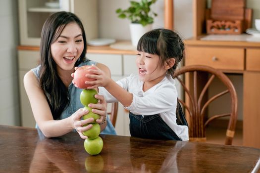 Asian mother excite and happy when her little girl put red apple over stack of green apple on table in the kitchen.