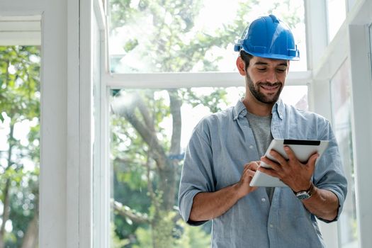 Caucasian engineer man with blue hardhat use tablet in office with glass window and he look happy with work.