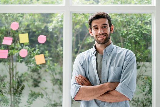 Business Caucasian man stand with arm crossed and confidence action with smiling look at camera in office with grass window and some note as background.