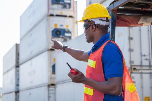 African American foreman or cargo container worker hold walkie talkie and point to front direction look like communicate with his team to work in workplace.
