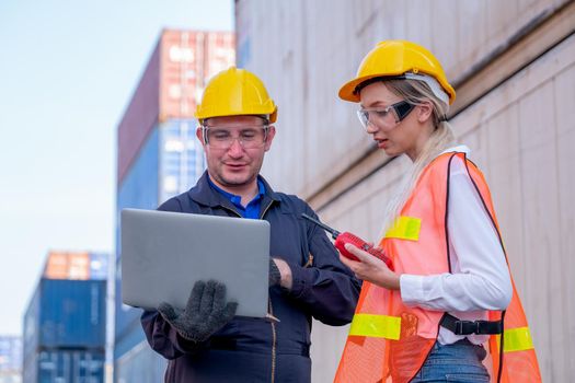 Two foreman and cargo container worker discuss together in workplace area with one hold and using laptop and the other hold walkie-talkie to contact their team.