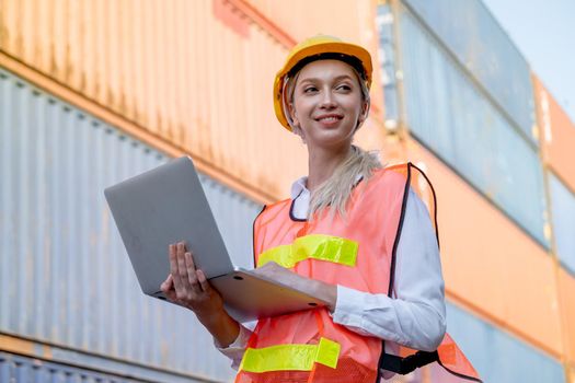 Beautiful foreman woman hold laptop and smile during working in cargo container workplace with background container stack with multi-color. Industrial system support employee and staff work concept.